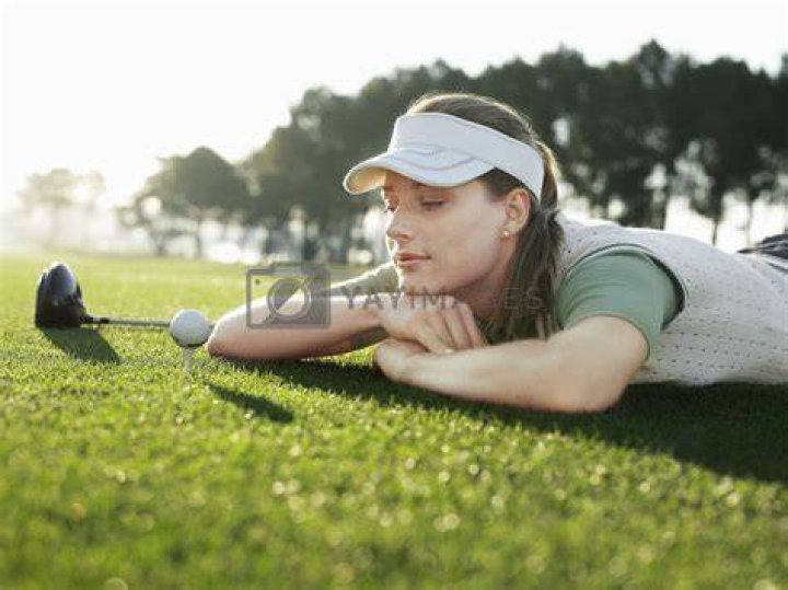 young female golfer lying on green, holding ball in front of eye, view from above